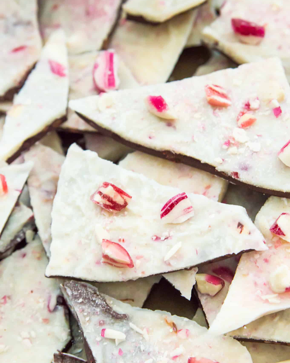 vegan white chocolate peppermint bark with two layers of chocolate and vegan candy canes crumbled on top. the bark is broken into shards and shown on a pile