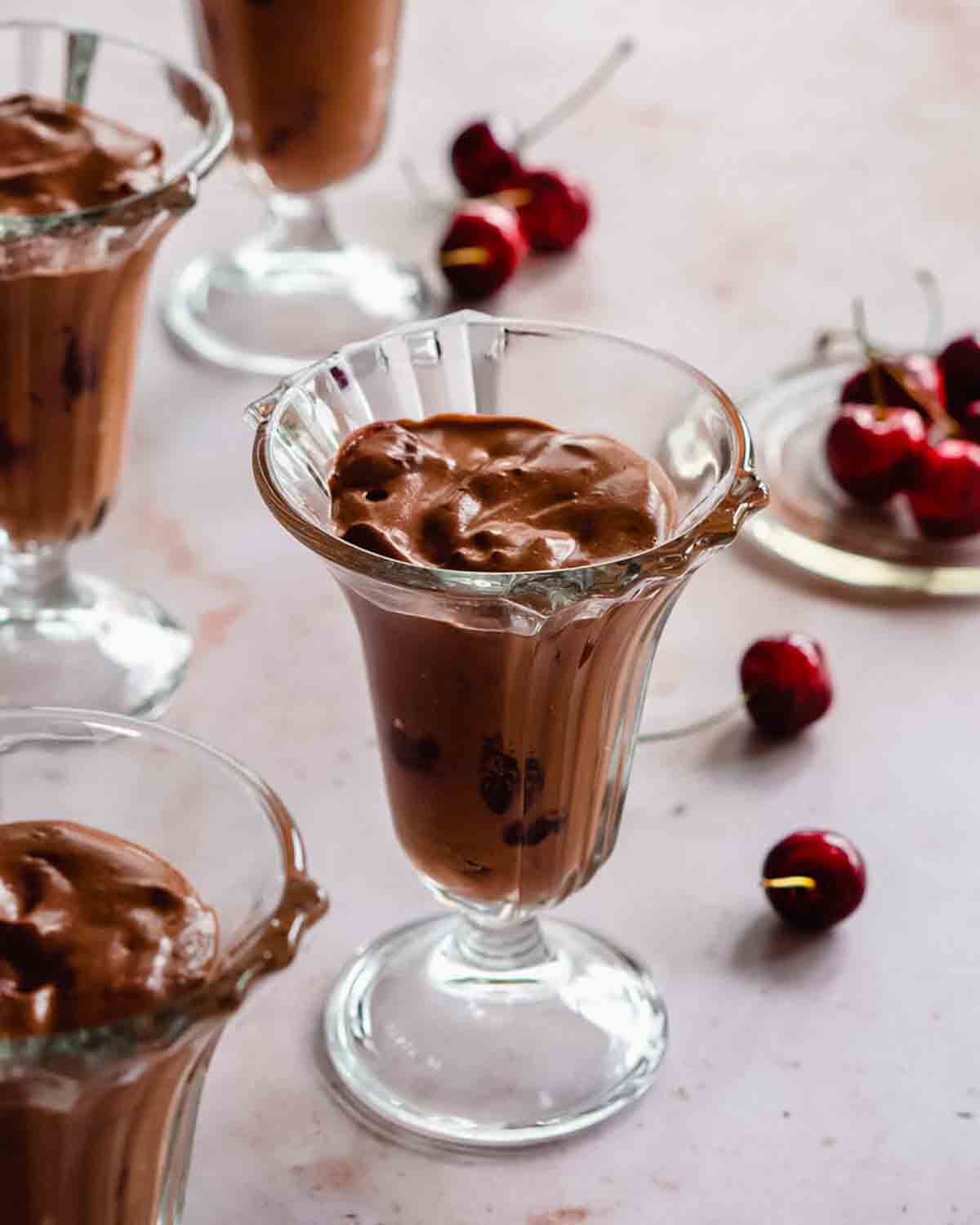 vegan tofu chocolate mousse in a beautiful dessert jar, surrounded by fresh cherries and multiple dessert jars visible in the foreground and background