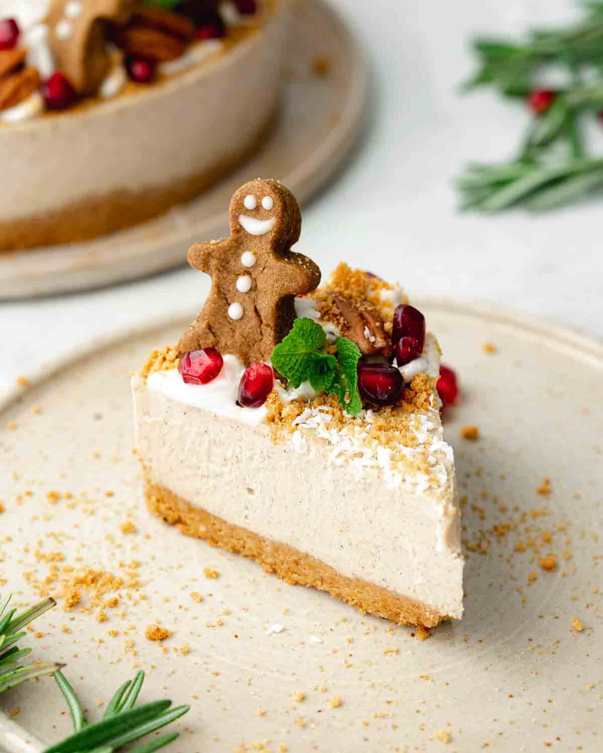 a slice of vegan gingerbread cheesecake on a white plate, decorated with a vegan gingerbread man and pomegranate seeds on top