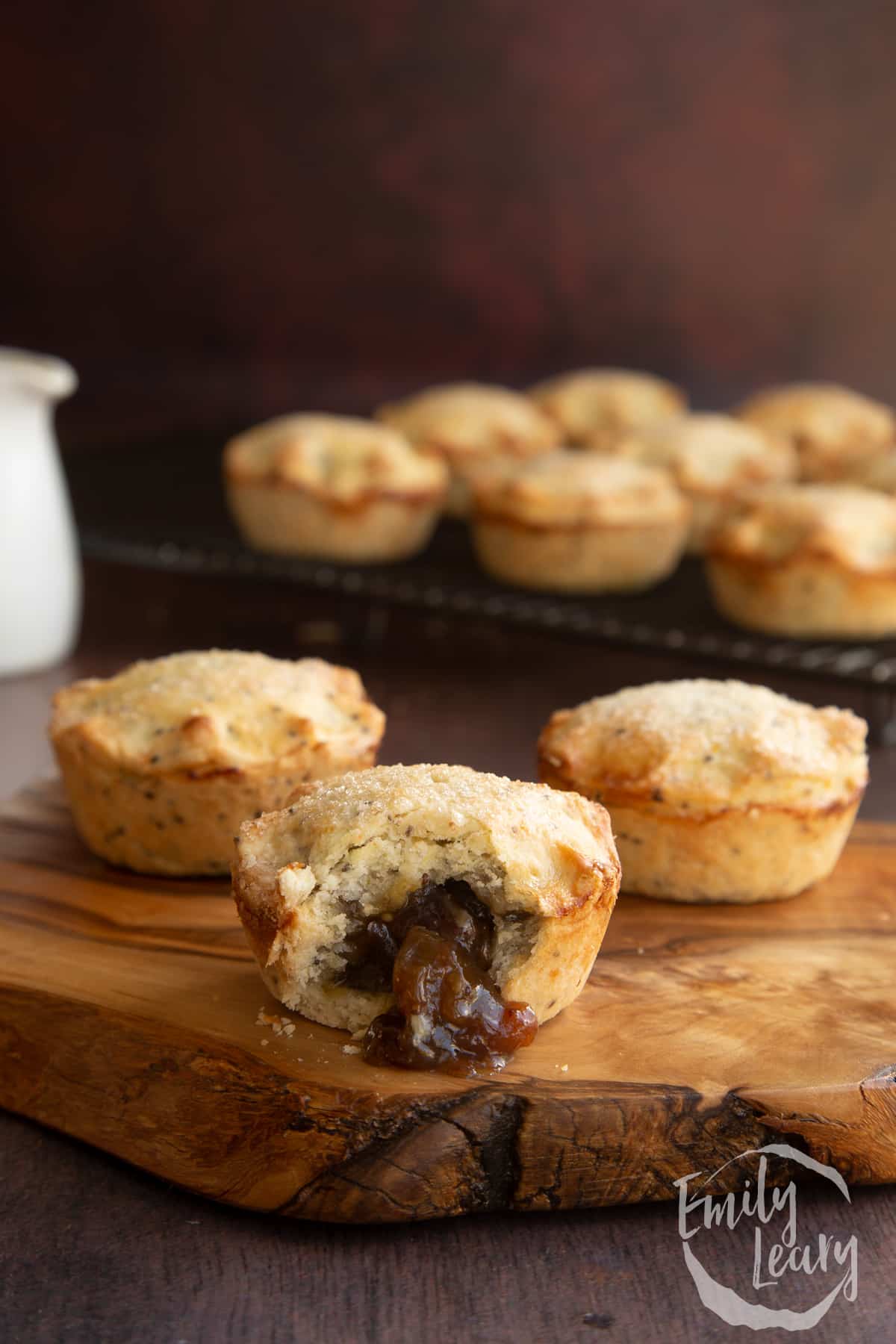 small vegan mince pies sitting on a wooden board with a bite taken out of the mince pie in the front. More vegan mince pies visible on a cooling rack in the background.