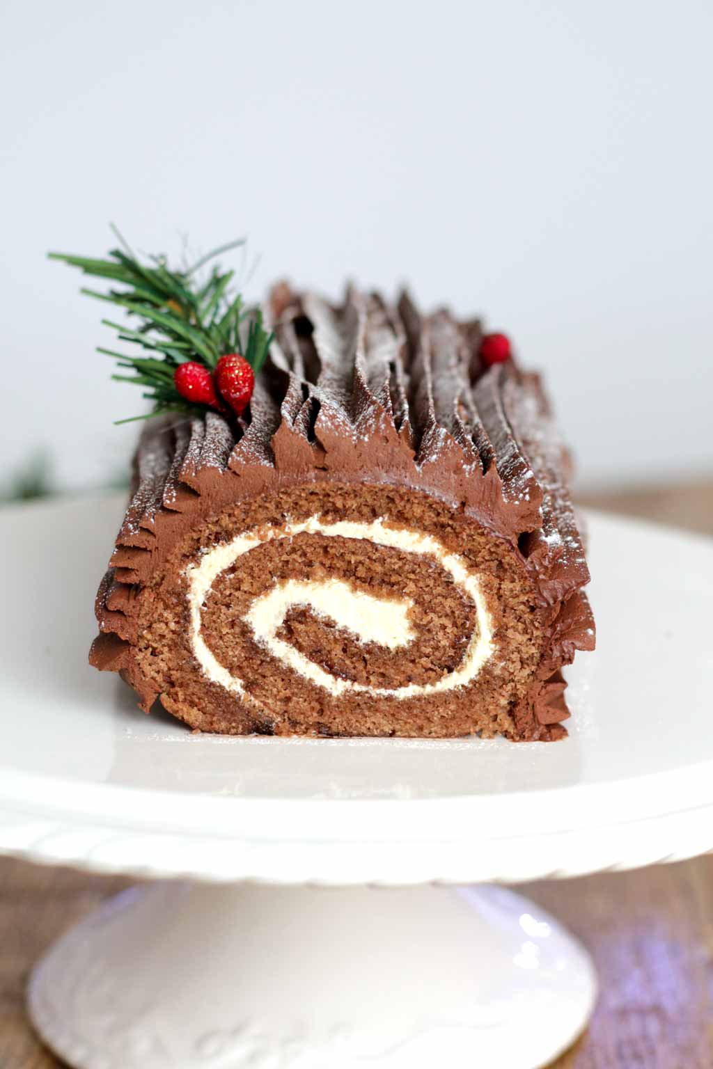 a vegan chocolate yule log on a white cake stand, decorated with cranberries and greenery on top. seen from the front with the rolled layers of the yule log clearly visible. the log is chocolate cake filled with a white vegan buttercream