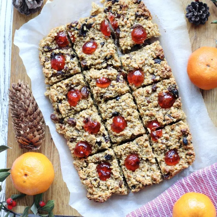 vegan mincemeat flapjacks as seen from above, they are sliced, on top of white parchment paper and surrounded by ingredients, such as oranges, The flapjacks have their fruit and nuts visible from the top