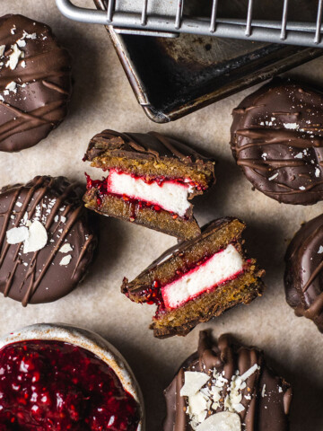 vegan wagon wheels on parchment paper, coated in chocolate and one wagon wheel cut in half in the middle showing the layers of biscuit, chocolate, marshmallow and raspberry chia jam