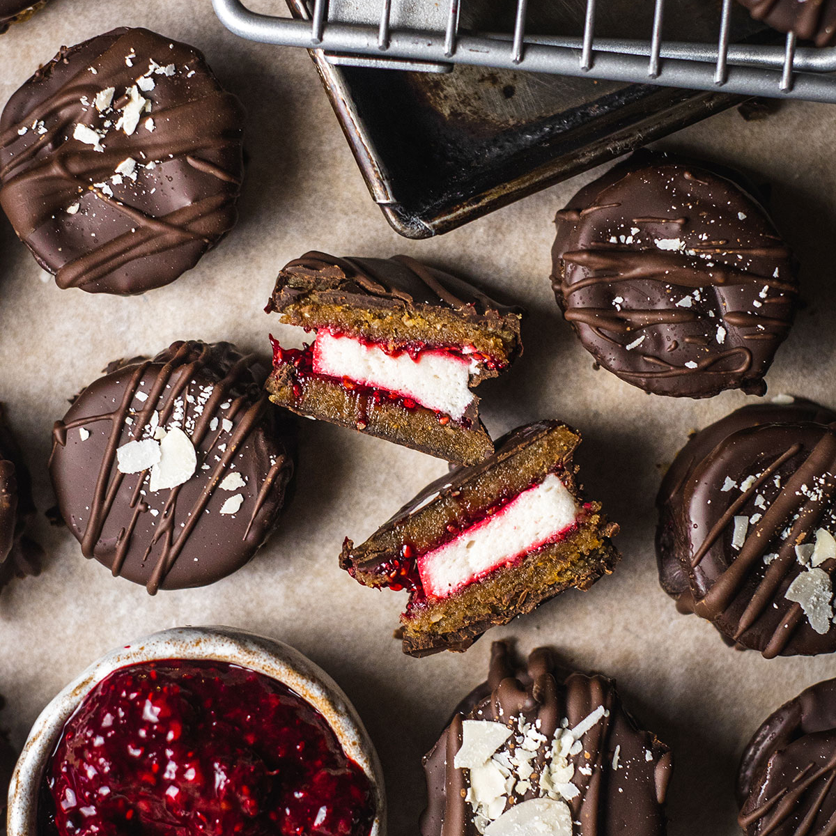 vegan wagon wheels on parchment paper, coated in chocolate and one wagon wheel cut in half in the middle showing the layers of biscuit, chocolate, marshmallow and raspberry chia jam