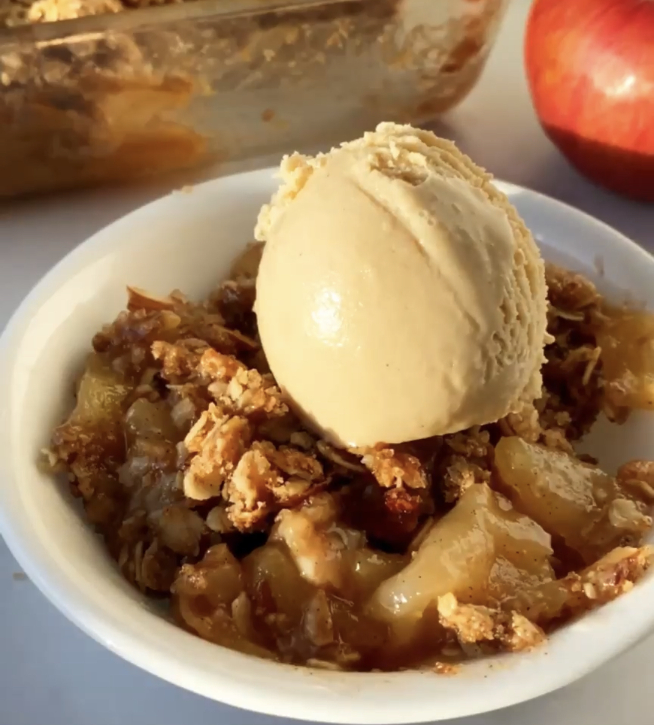 a white bowl filled with vegan apple crumble dessert and a dollop of vegan ice cream on top
