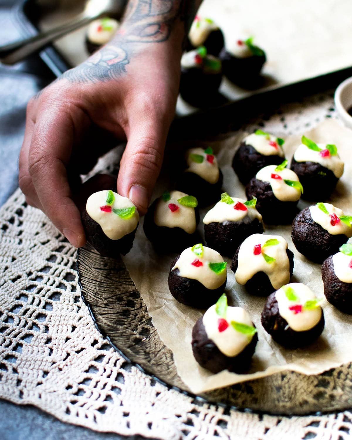 vegan mini christmas pudding truffles on a platter with a hand coming in to lift one off the platter. all the truffles are small and round and decorated like a christmas pudding with white frosting on top