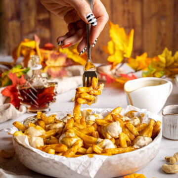 vegan poutine in a serving dish with a hand and fork lifting off some french fries from the top