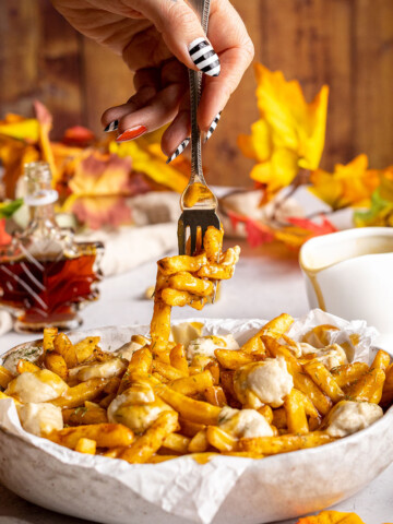 vegan poutine in a serving dish with a hand and fork lifting off some french fries from the top