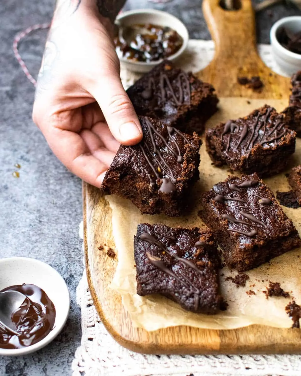 vegan christmas brownies on a wooden board with a parchment paper underneath. a hand is coming in to grab one of the brownies and ingredients are surrounding the board