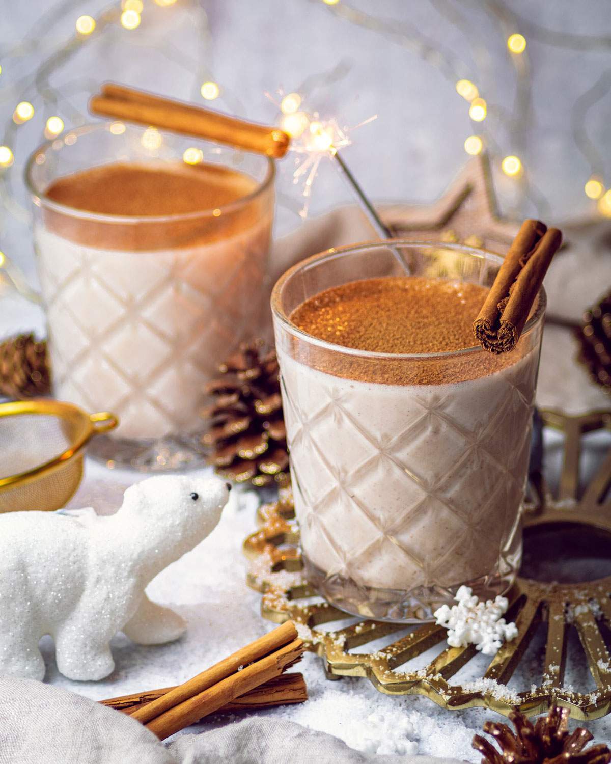 2 glasses of oat milk eggnog are sitting on a festive table setup with a cute polar bear decoration.