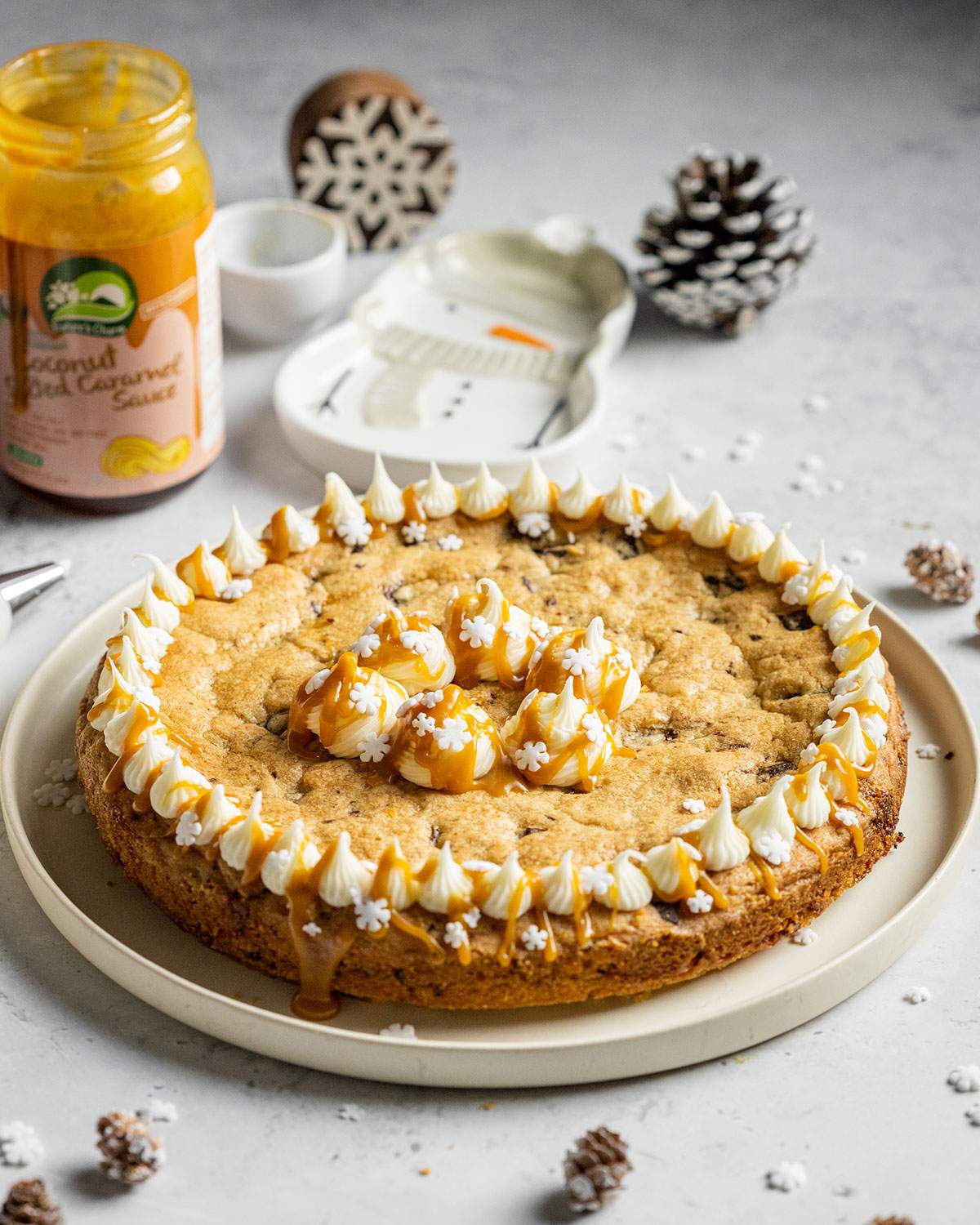 a round vegan cookie cake topped with vegan frosting and salted caramel sauce on a white table next to a jar of vegan caramel sauce