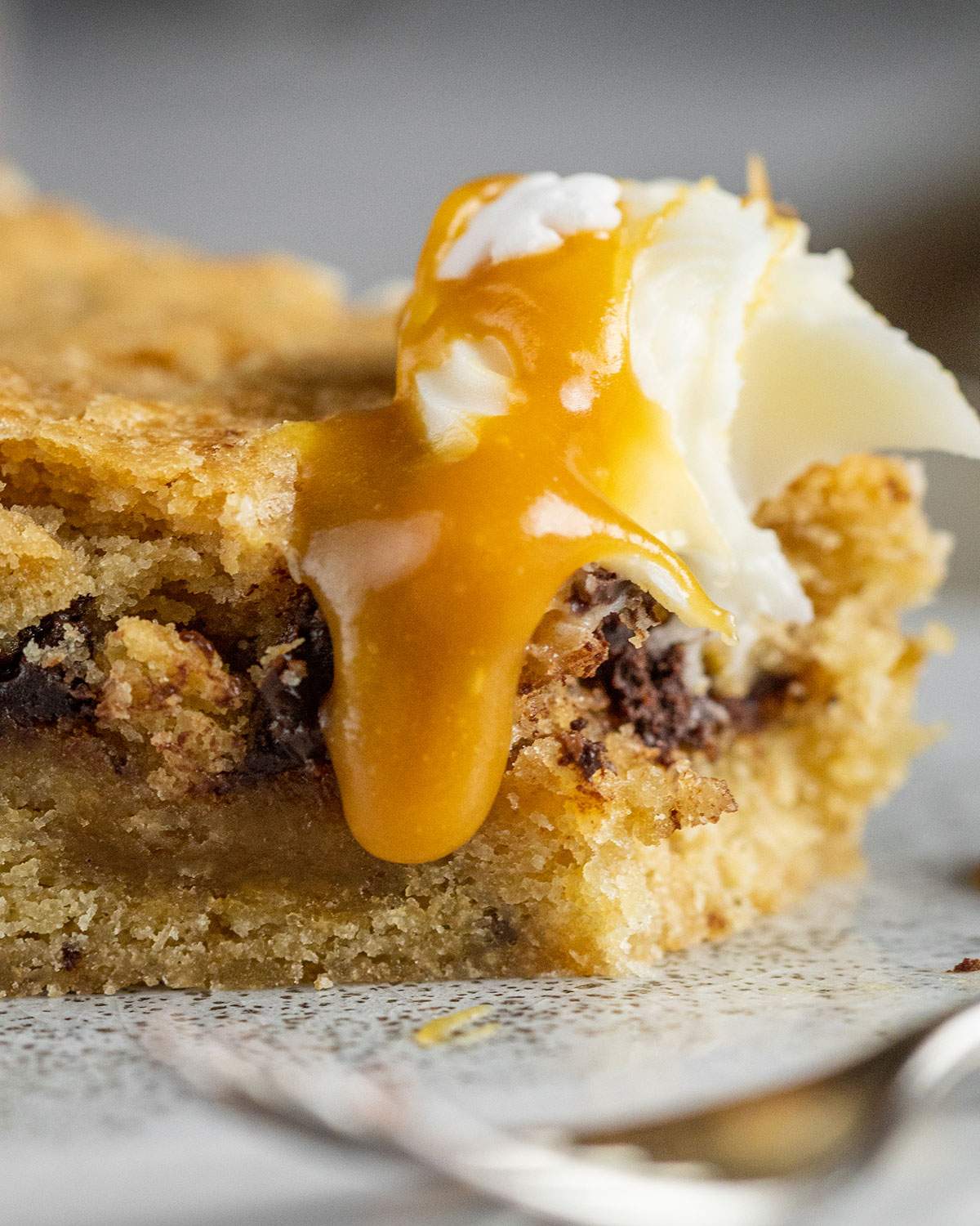 a slice of vegan chocolate chip cookie cake with vegan frosting and vegan caramel sauce with a piece being taken off the tip of the slice