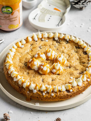 a round vegan cookie cake topped with vegan frosting and salted caramel sauce on a white table next to a jar of vegan caramel sauce