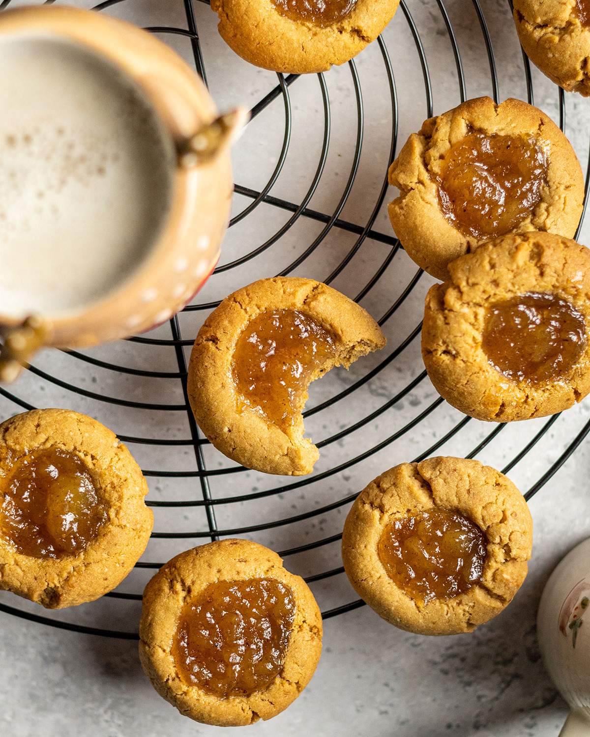 a cooling rack covered with vegan thumbprint cookies and a cosy mug, one cookie has a bite taken out of it