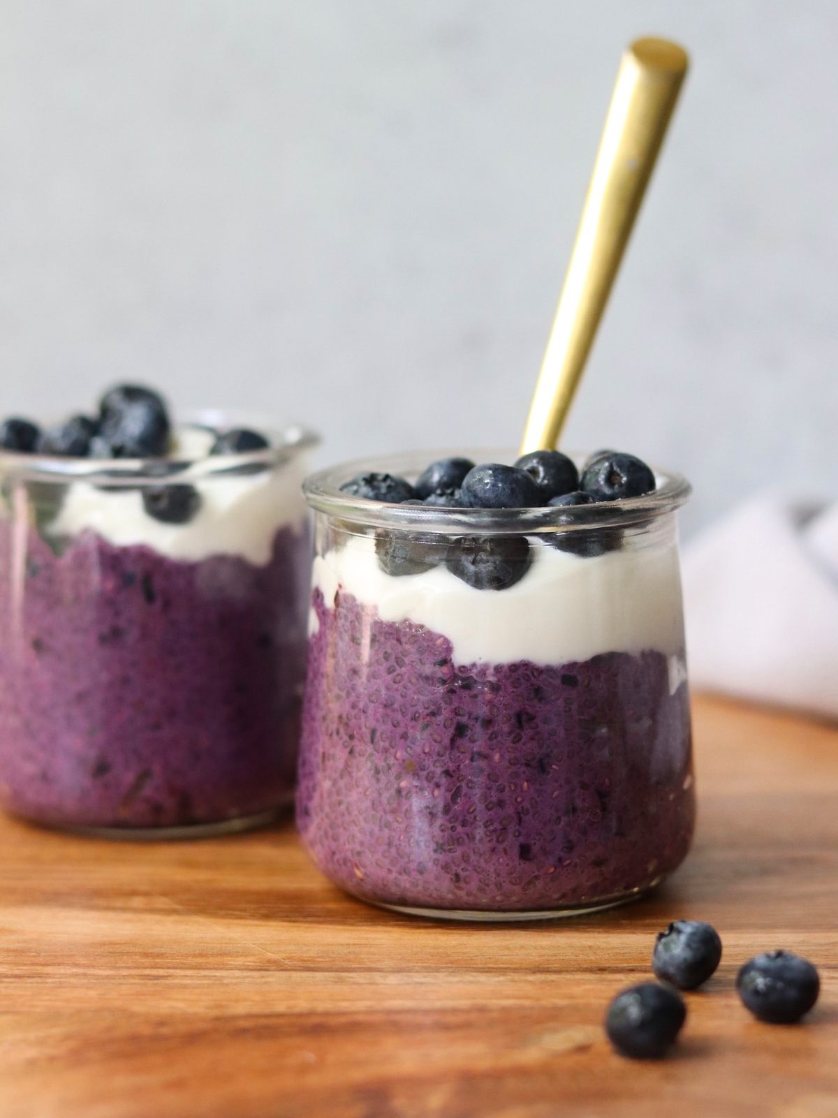 two glass jars of blueberry chia pudding on a wooden table, a golden spoon coming out of one of them