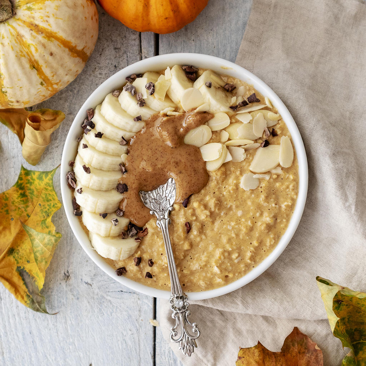a bowl filled with pumpkin spice porridge and topped with fresh fruits such as banana, nuts, cacao nibs and nut butter. A spoon is sitting on top of the bowl.