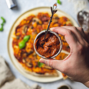 a small pot with homemade roasted tomato pizza sauce held by a hand over a pizza