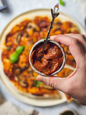 a small pot with homemade roasted tomato pizza sauce held by a hand over a pizza