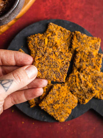 a hand holding a homemade sweet potato cracker next to a vegan cheeseboard