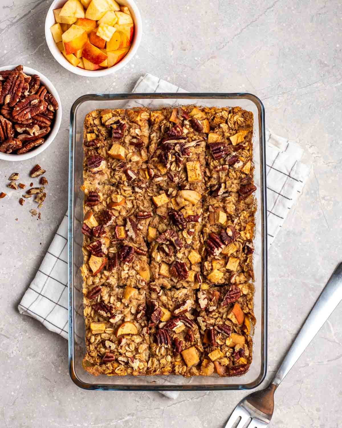 a glass dish filled with vegan apple oat meal on a table with pecans scattered across the top and a bowl of apples and pecans on the side