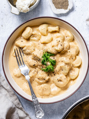 a white bowl filled with vegan tortellini and a creamy cauliflower alfredo sauce with pepper and fresh herbs on top