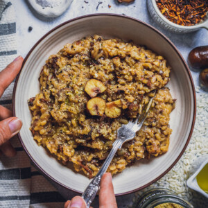 a bowl filled with vegan mushroom risotto and topped with chestnuts. Theres hands next holding the bowl and a fork grabbing some risotto