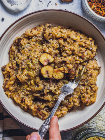 a bowl filled with vegan mushroom risotto and topped with chestnuts. Theres hands next holding the bowl and a fork grabbing some risotto