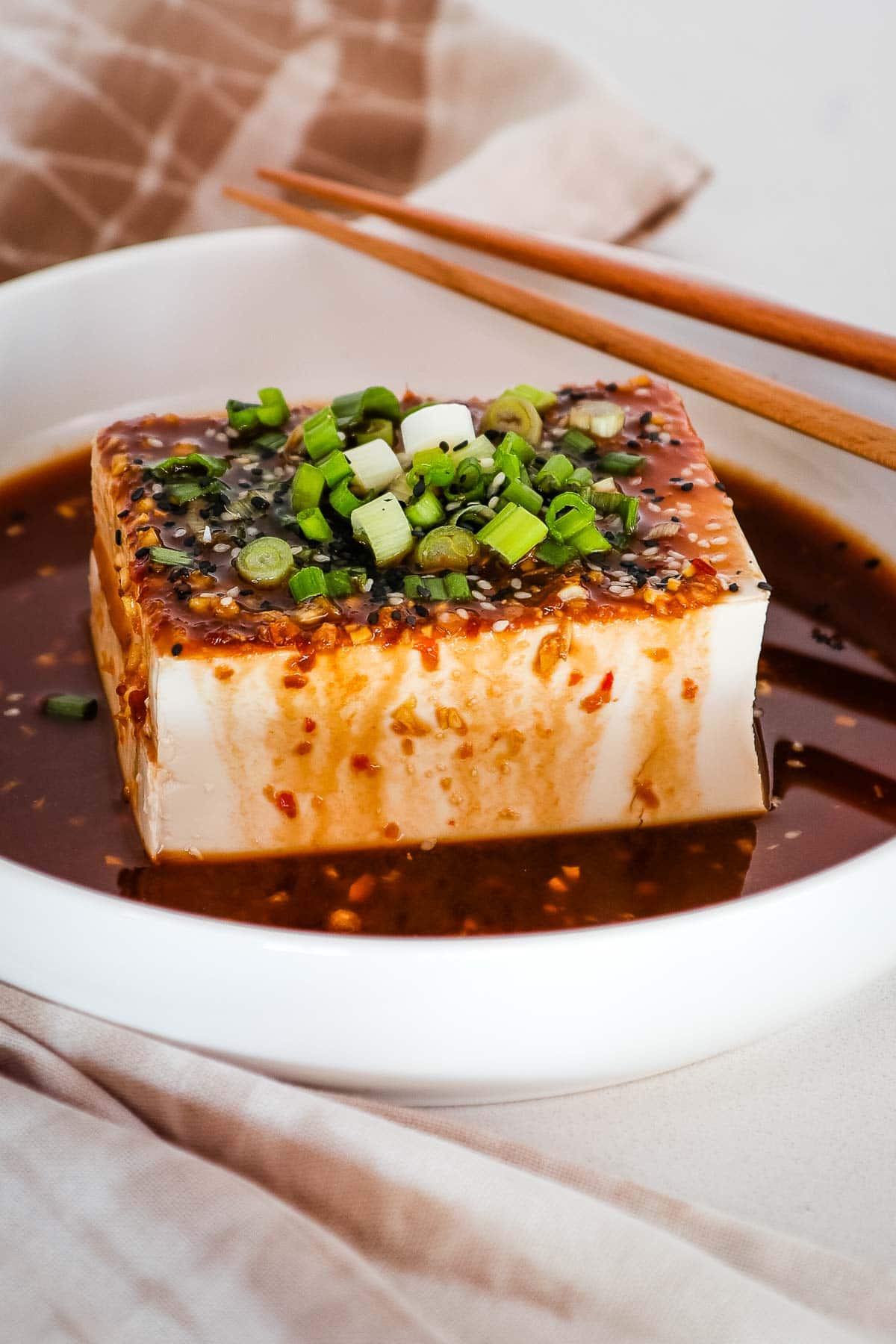 A white rectangular plate filled with silken tofu cubes, drizzled with a spicy soy sauce and garnished with chopped green onions. The dish is a protein-rich and flavorful vegan appetizer or side dish.