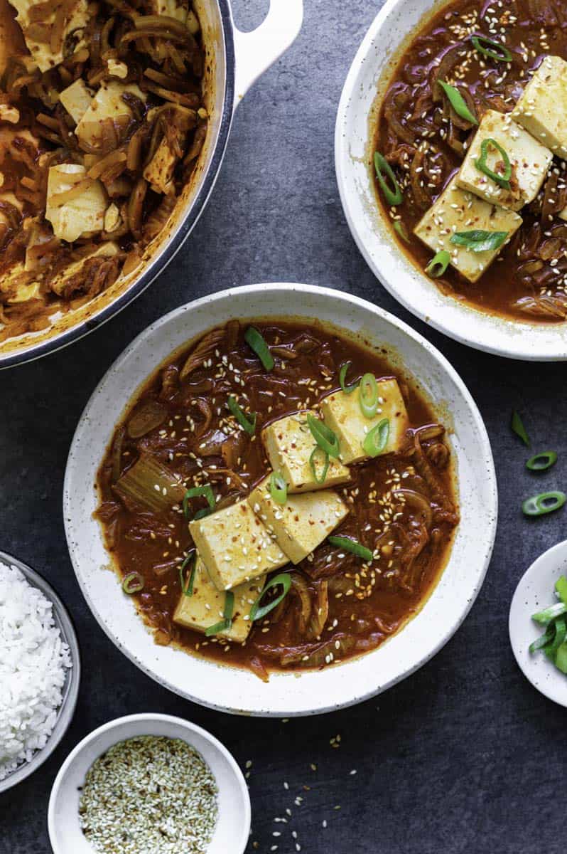 A steaming bowl of vegetarian kimchi jjigae, a spicy Korean tofu stew filled with silken tofu, kimchi, and vegetables. The stew is garnished with green onions and red pepper flakes, adding a pop of color and spice to the dish.