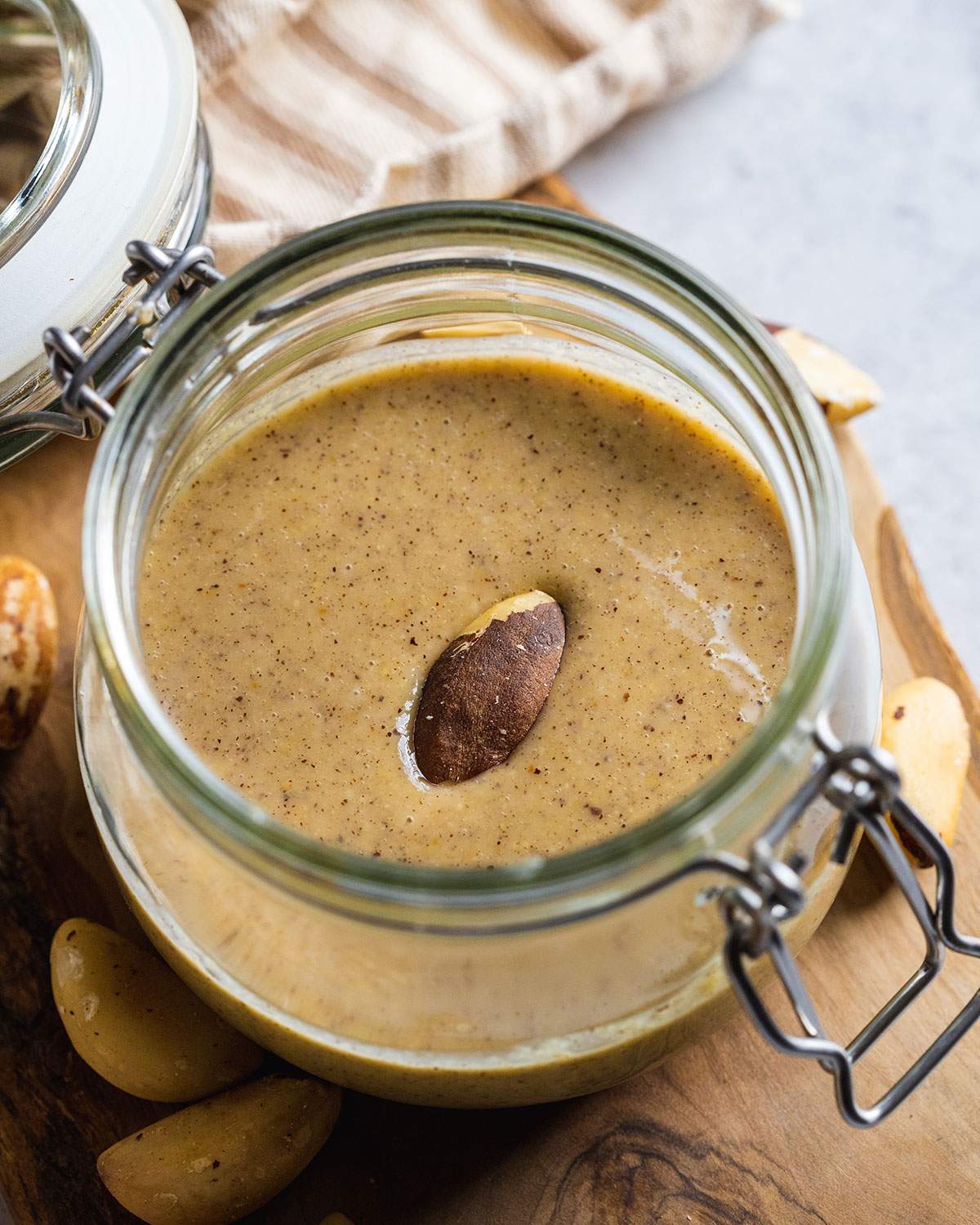 brazil nut butter in a glass jar on a wooden tray with raw nuts around and one nut in the middle of the nut butter