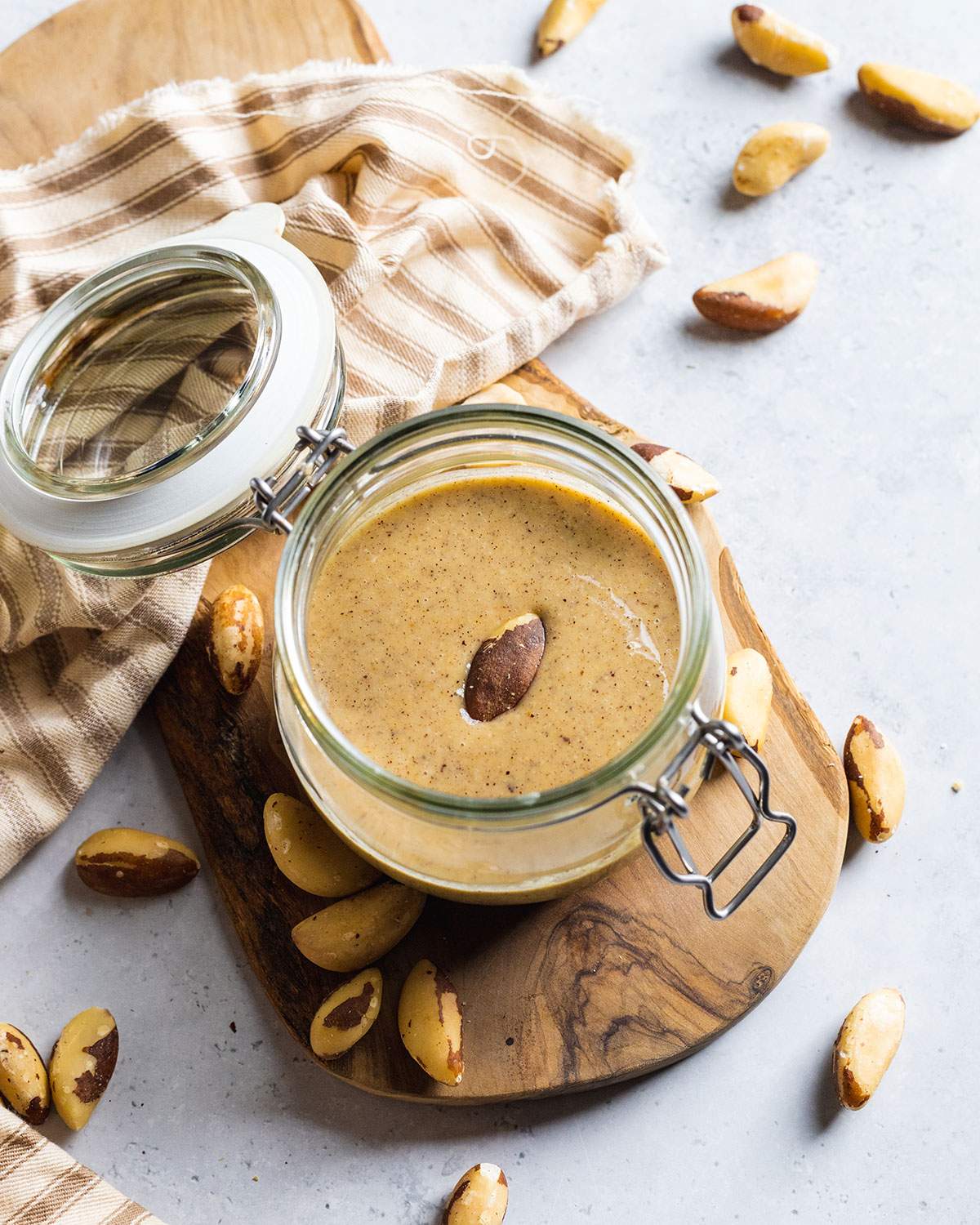 brazil nut butter in a glass jar on a wooden tray with raw nuts around and one nut in the middle of the nut butter