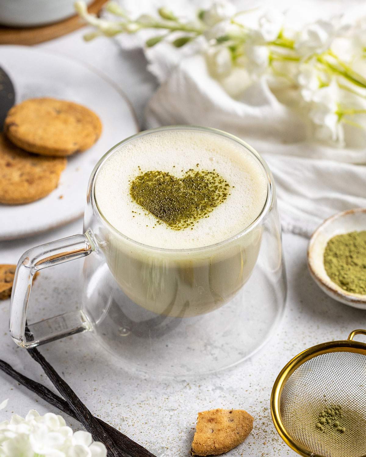 frothy vanilla matcha latte with a matcha heart in the middle on a white table with cookies next to it