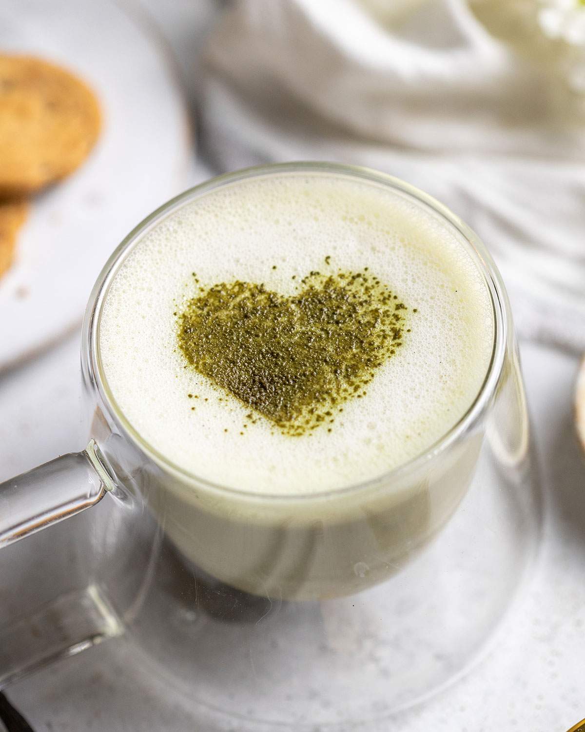 frothy vanilla matcha latte with a matcha heart in the middle on a white table with cookies next to it