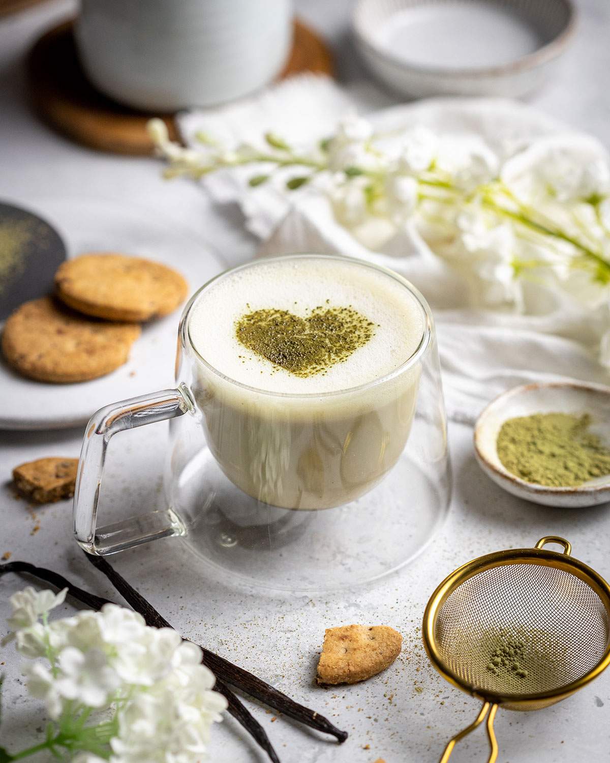 frothy vanilla matcha latte with a matcha heart in the middle on a white table with cookies next to it