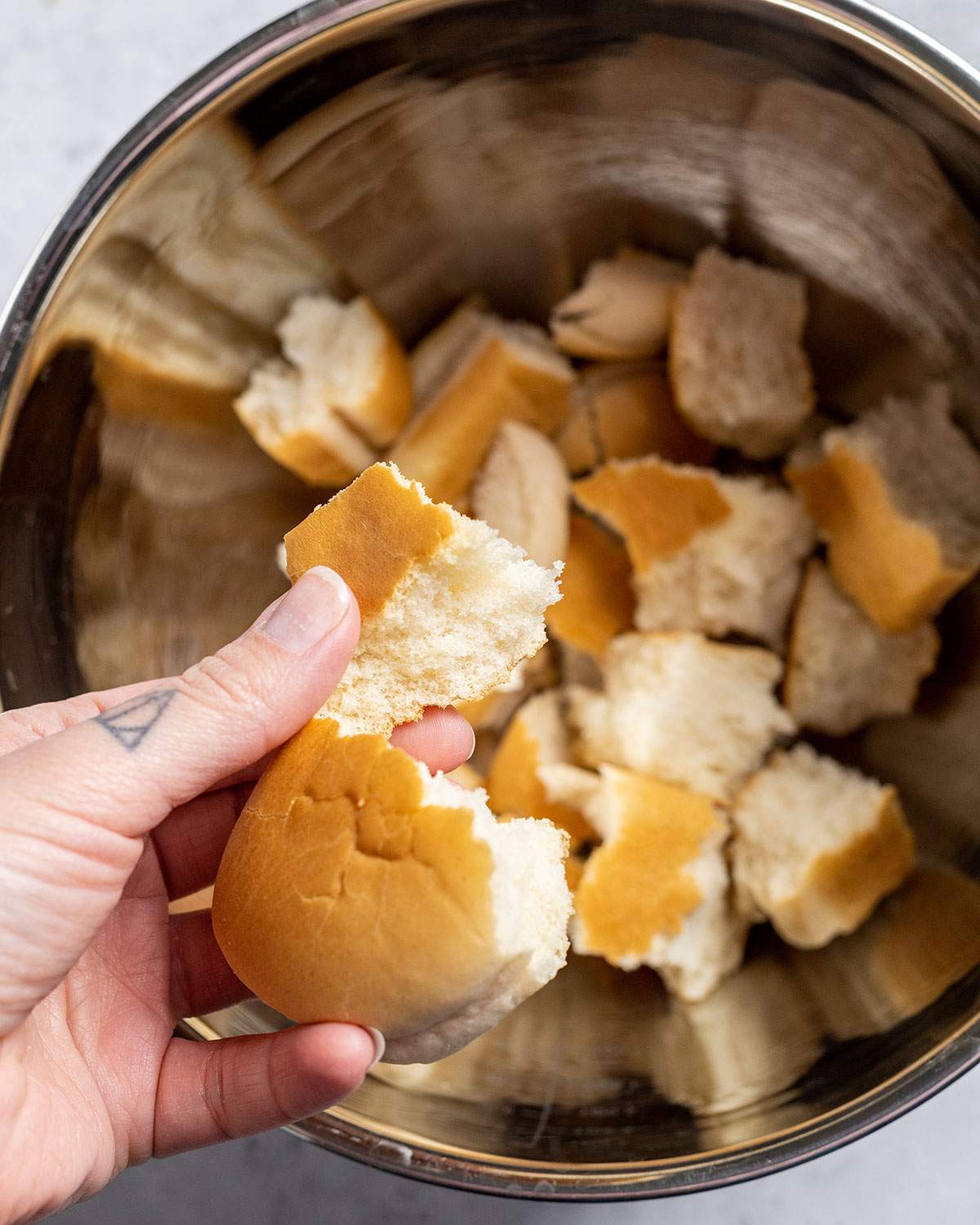 A hand ripping vegan bread rolls into a large mixing bowl.