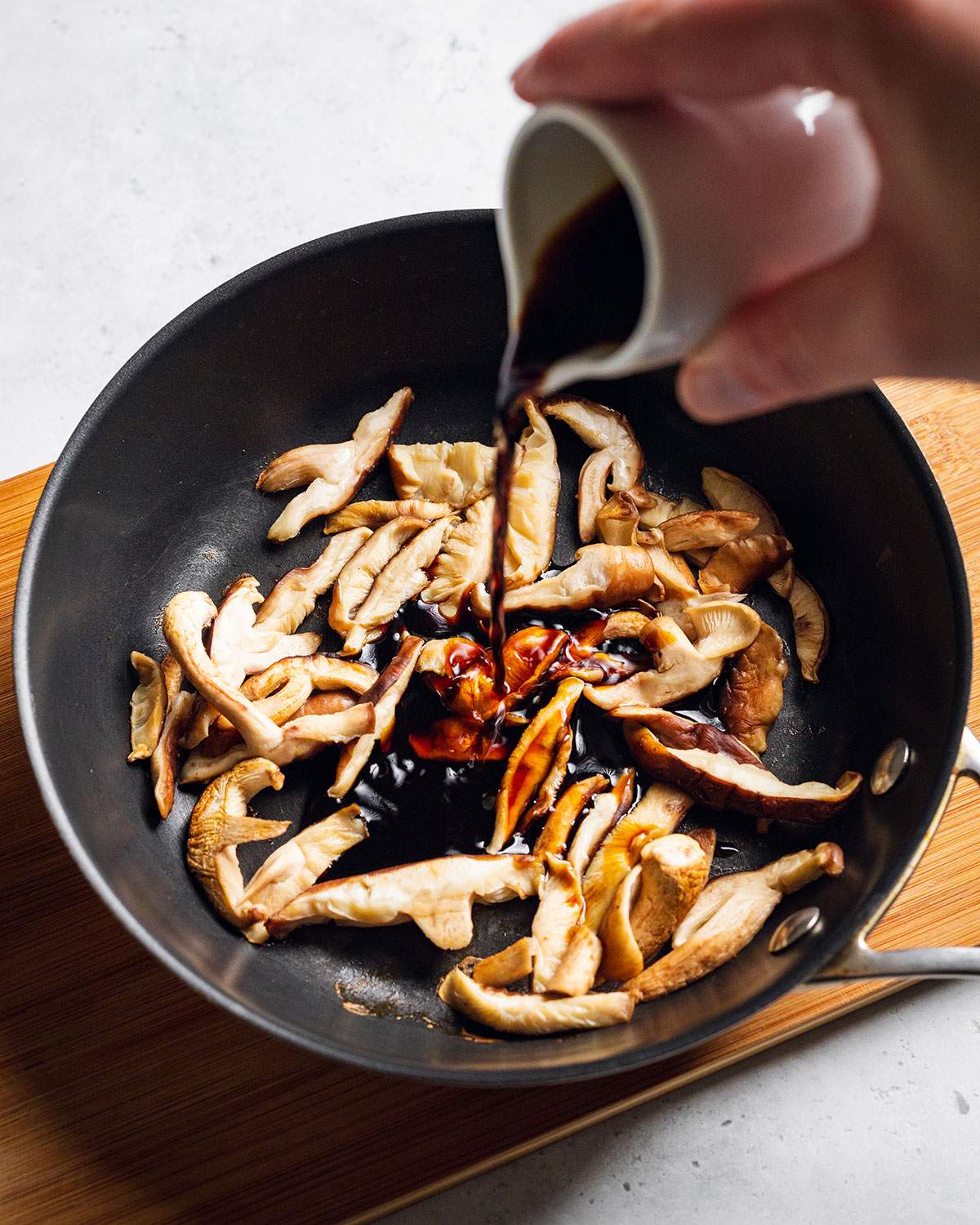 Pouring tamari sauce onto sliced shiitake mushrooms in a pan.