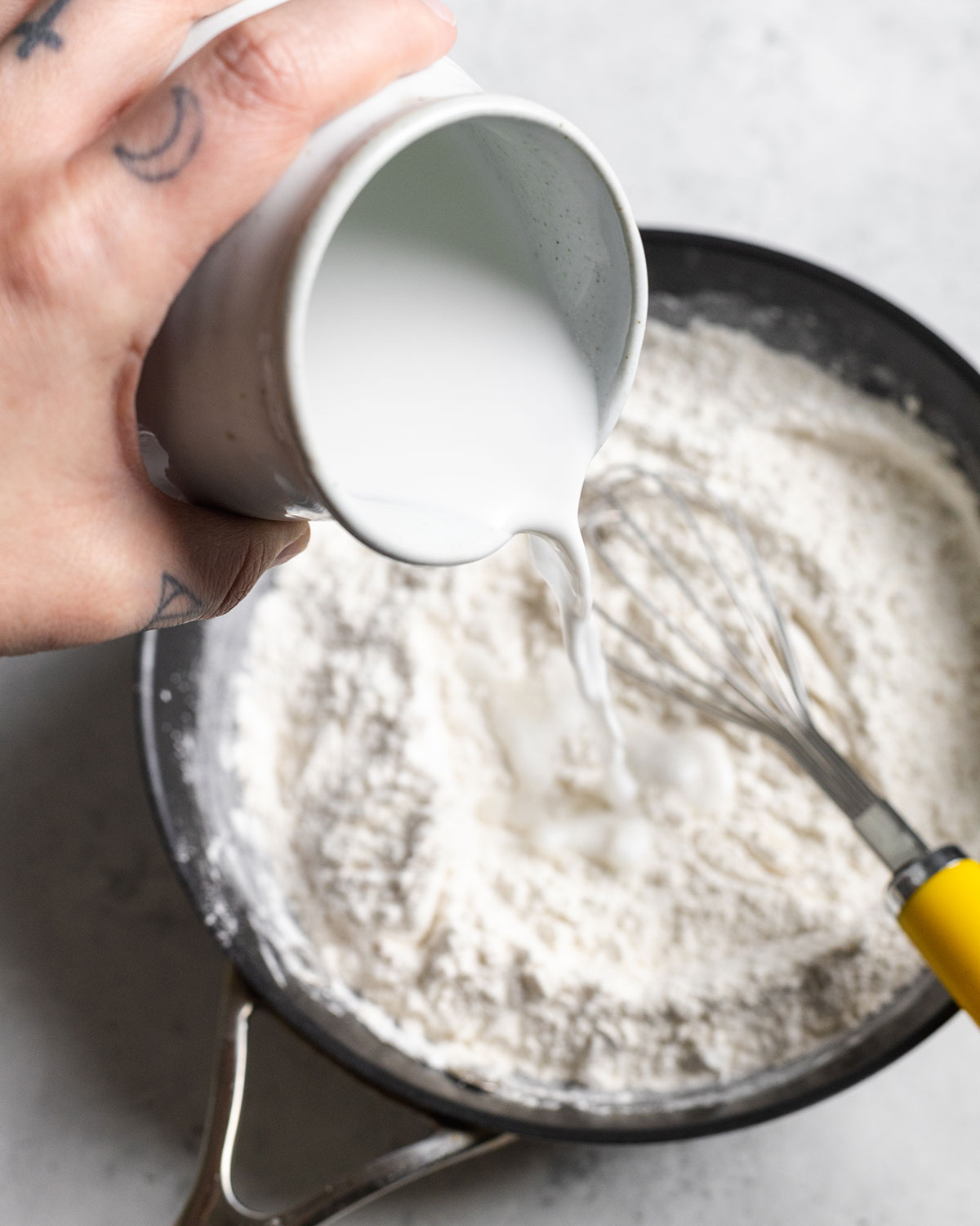 A hand pouring coconut milk from a jug into the dry ingredients in a pan.