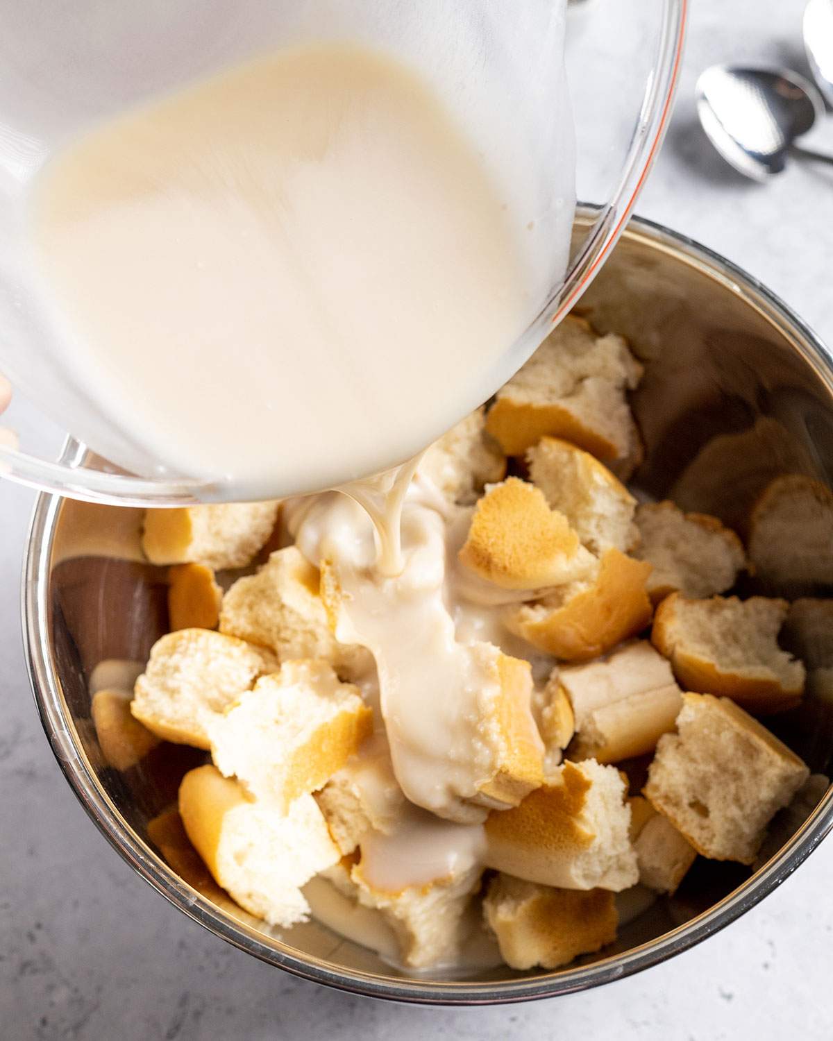 Pouring a mix of vegan condensed milk and evaporated coconut milk onto stale bread in a mixing bowl.