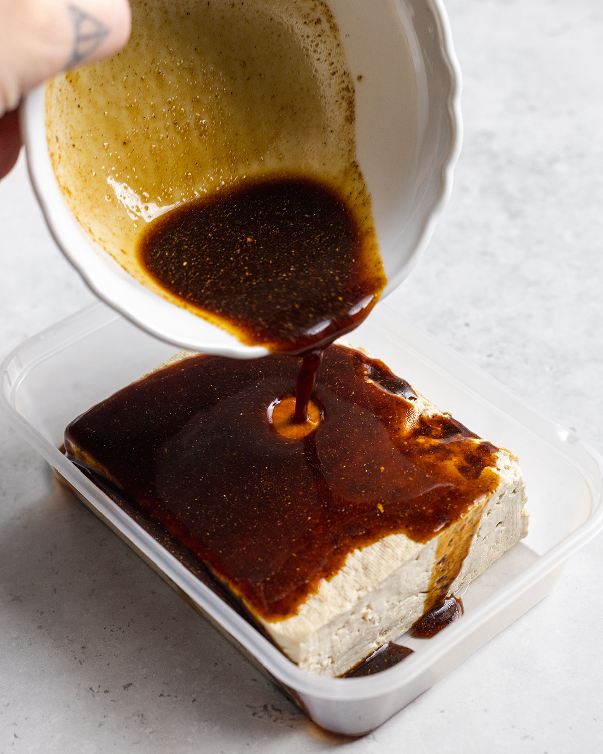 Pork Belly marinade dripping from a white mixing bowl onto a block of pressed tofu in a fridge container.