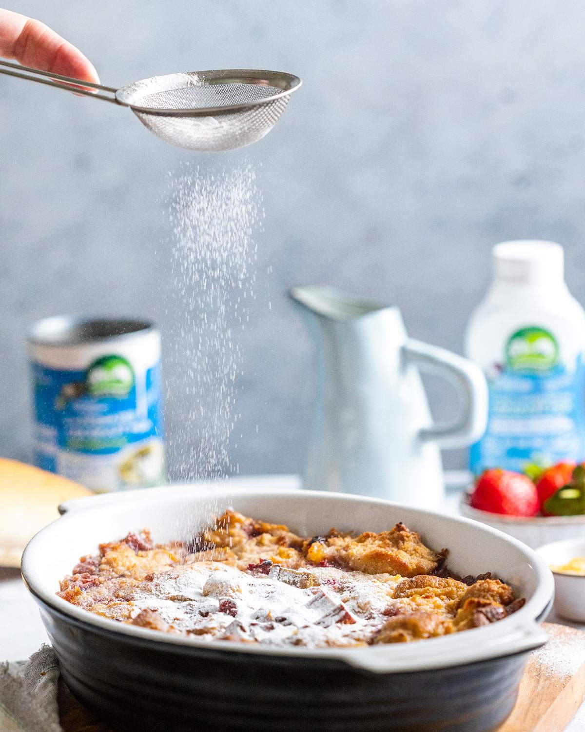 A hand sprinkling powdered sugar onto baked bread pudding using a fine sieve.