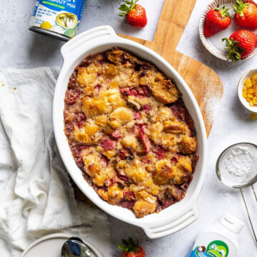 An oven dish filled with baked vegan bread pudding with strawberries and rhubarb on a wooden serving board next to ingredients.