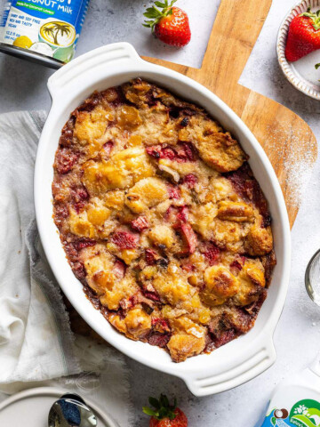 An oven dish filled with baked vegan bread pudding with strawberries and rhubarb on a wooden serving board next to ingredients.