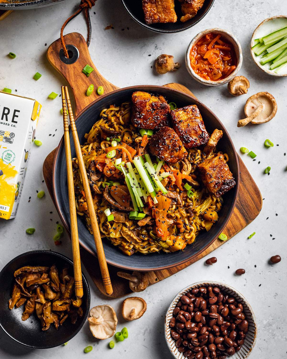 A bowl of plant-based miso ramen with black beans on a wooden board with ingredients around it.