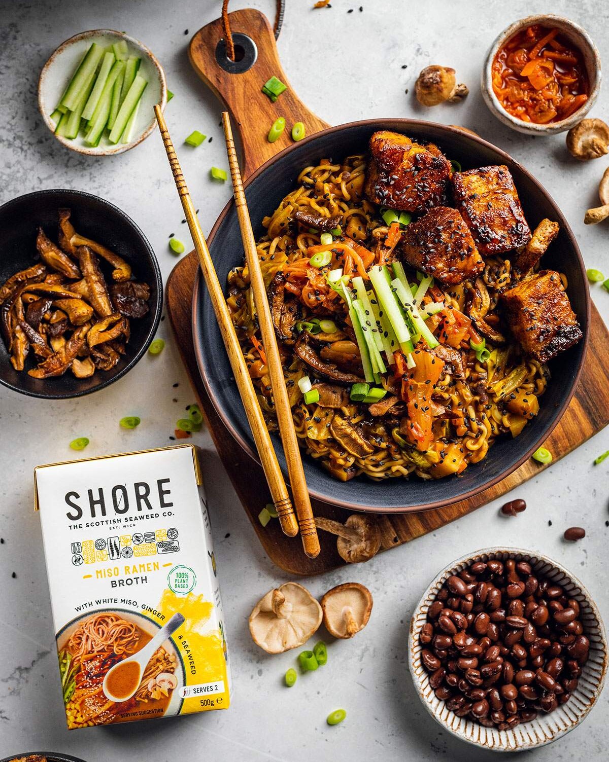 A bowl of vegan ramen with black beans and tofu pork belly on a white table with ingredients surrounding the bowl.