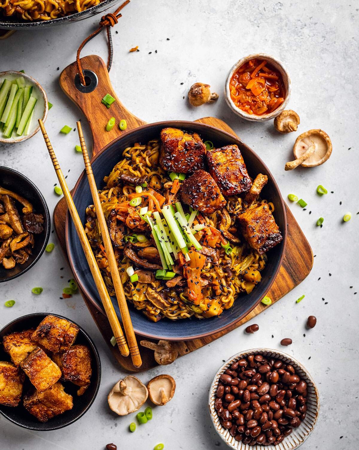 A bowl of vegan ramen with black beans and tofu pork belly on a white table with ingredients surrounding the bowl.