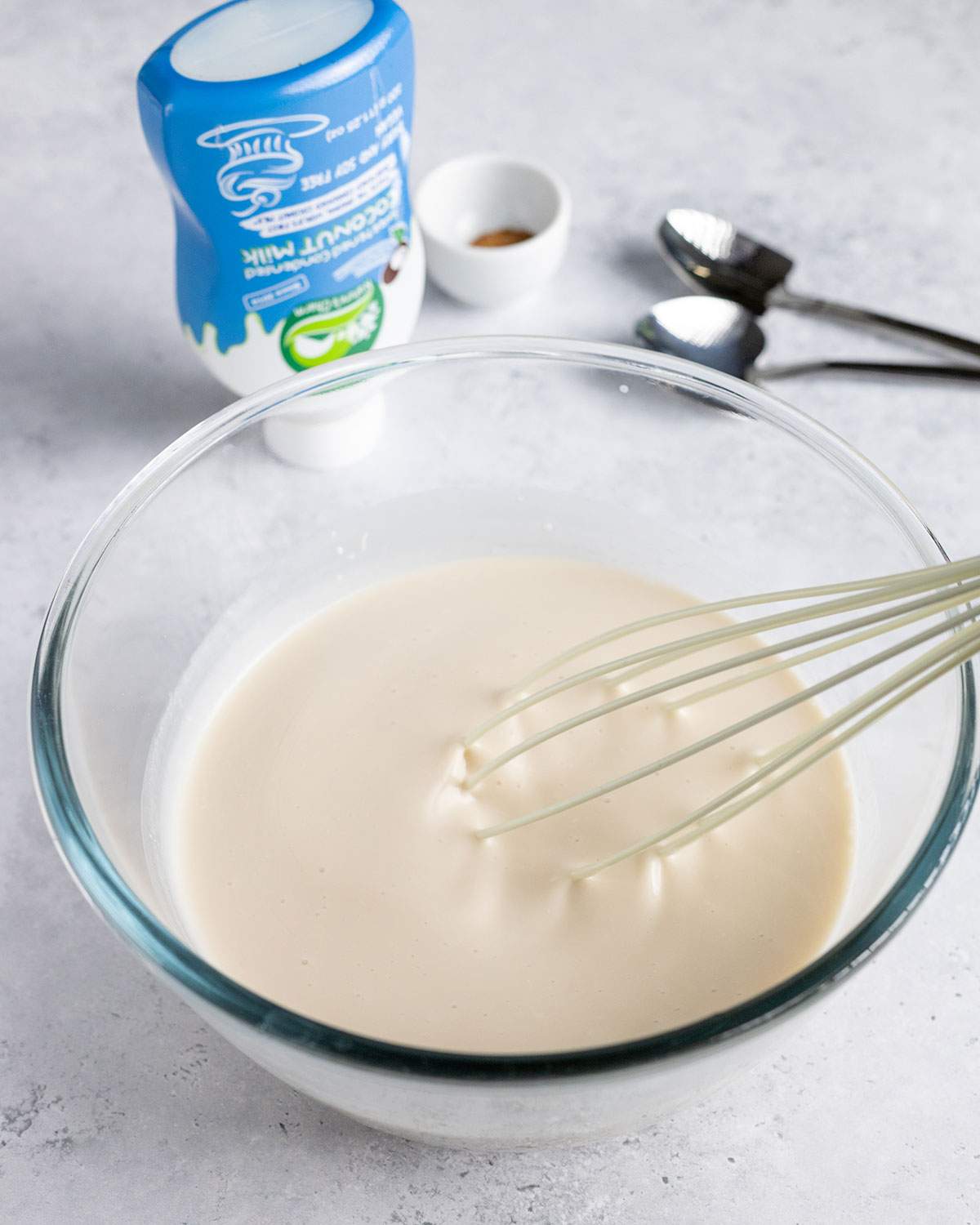 A whisk mixing vegan condensed coconut milk and evaporated milk in a glass bowl.