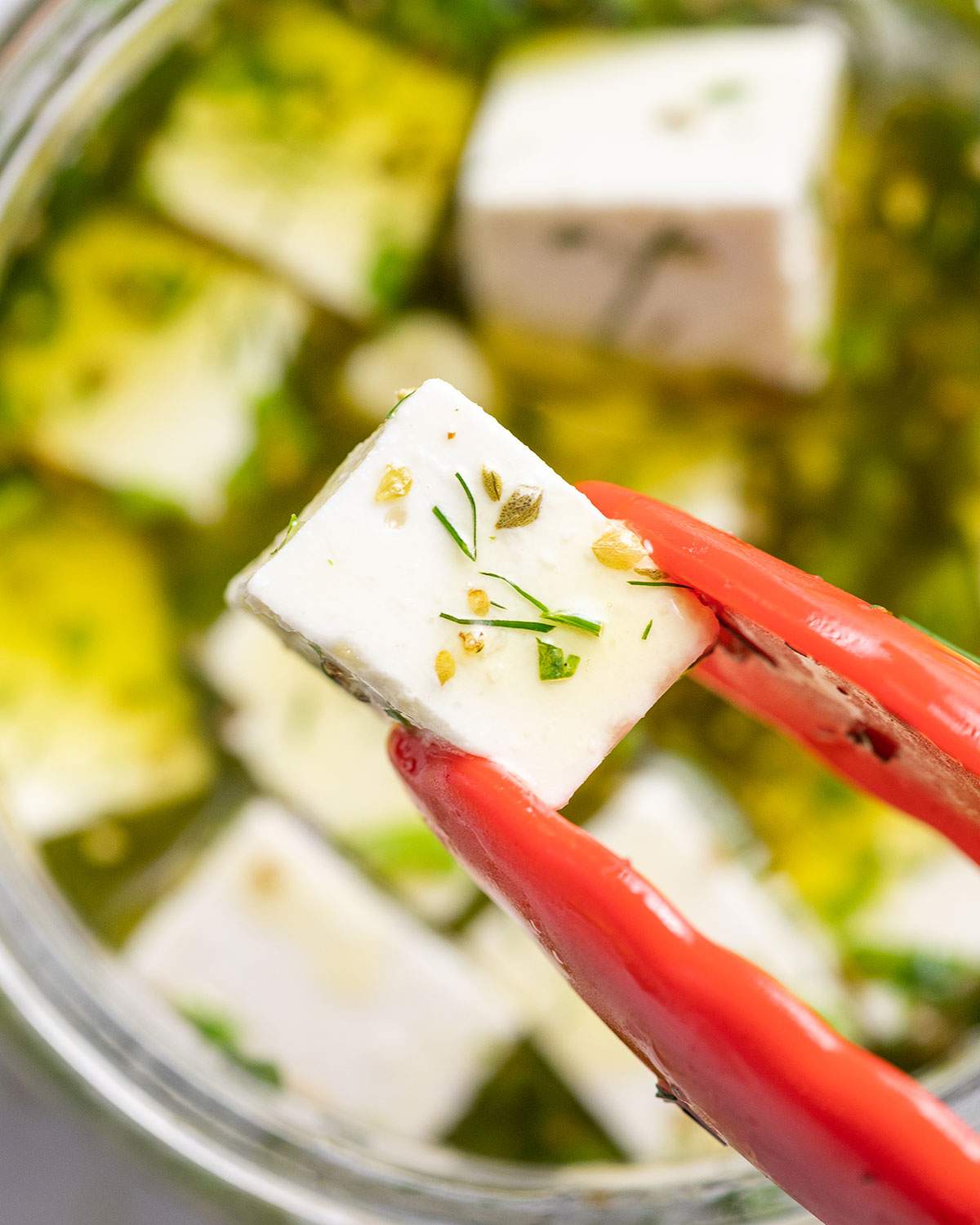 A cube of vegan feta held up close above a jar of more feta cubes in marinade.