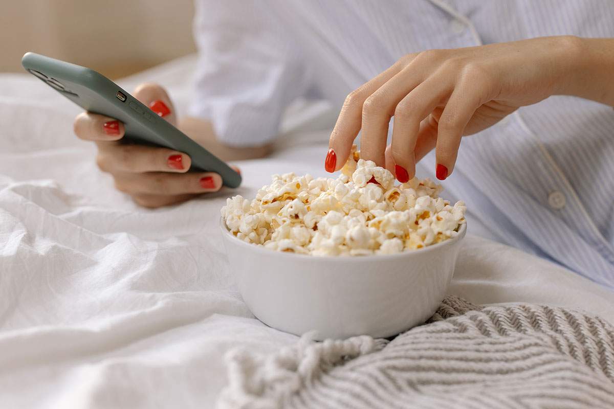 A hand reaching for a bowl of popcorn on a blanket, whilst the person holds a phone in the other hand.