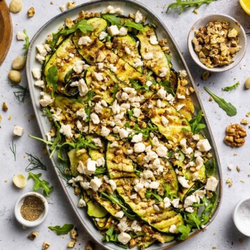 Vegan Grilled Zucchini Salad with walnuts and vegan feta crumbles on a serving platter, sitting on a white dinner table surrounded by ingredients.