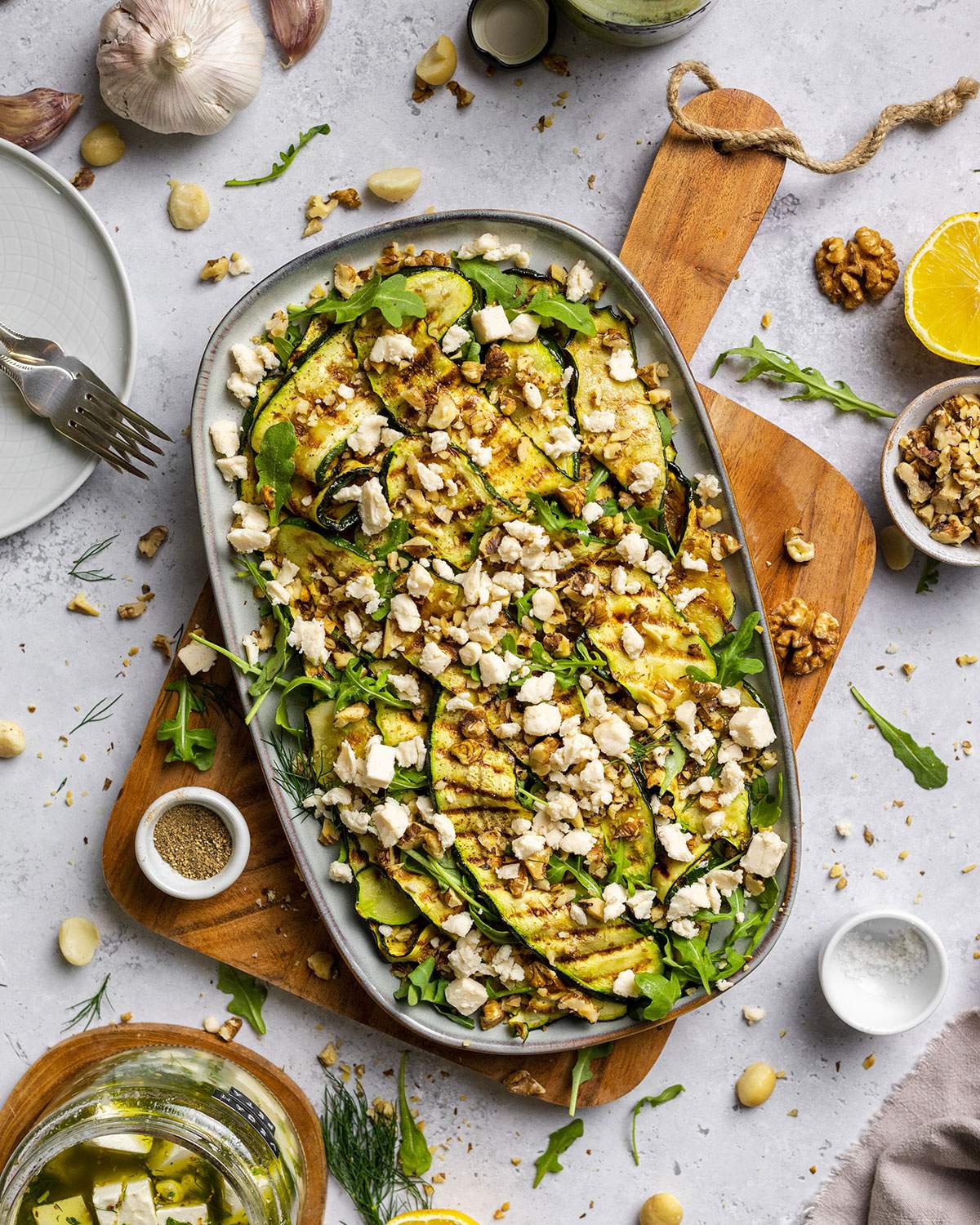 Vegan Grilled Zucchini Salad with walnuts and vegan feta crumbles on a serving platter, sitting on a white dinner table surrounded by ingredients.